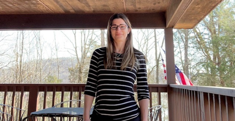 Beth is smiling and posing for the photo. She is outside on a wooden deck with a rail. Beyond the rail is a landscape of trees, mountains and sky.