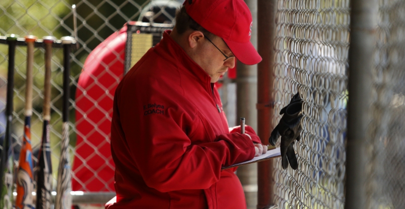 Ed is coaching his team from behind a chainlink fence and taking notes. Nearby is a rack of multi-color baseball bats.