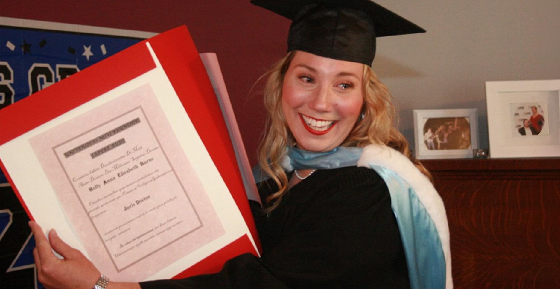 Holly is wearing a graduation gown and cap, holding her degree for the photograph. She is holding back tears and has a big joyful smile.