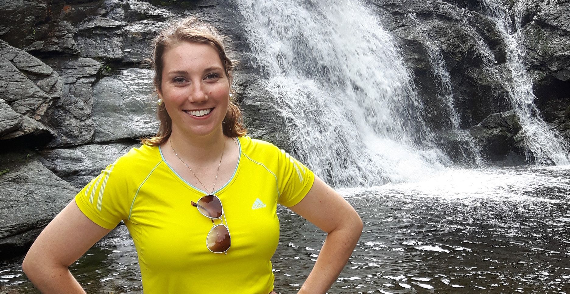 Holly is smiling and wearing a bright yellow shirt. She stands with her hands on her hips in front of a large waterfall.